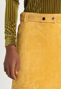 Jupe en suède jaune à la texture lisse, dotée d'une ceinture et de deux boutons noirs à la taille, associée à un haut côtelé vert.