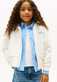 Biała bluza z zamkiem błyskawicznym nałożona na jasnoniebieską w paski koszulę z guzikami, zestawiona z białą koszulką i dżinsami. Osoba ma kręcone włosy i rękę w kieszeni.