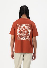 Tricou de culoare ruginiu, cu un design geometric �și floral alb pe spate. Mâneci scurte și o croială lejeră. Material din bumbac.