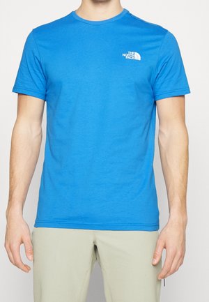 Mann trägt ein hellblaues kurzärmliges T-Shirt mit dem "The North Face"-Logo auf der linken Brust und beigefarbenen Hosen vor einem schlichten Hintergrund.