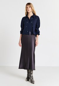 Donna che indossa una camicia blu navy con piccoli embellishments, una gonna midi grigio scuro lucida e stivali a metà polpaccio con motivo pitone, in piedi.