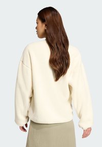 Krämfärgad fleece sweatshirt med sänkta axlar och långa ärmar. Har en mjuk textur och en avslappnad passform; matchas med en veckad beige kjol.