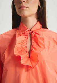 Blouse couleur corail avec un col froncé et une fermeture à patte. Tissu lisse et léger, présentant une ouverture en forme de clef au niveau du décolleté.