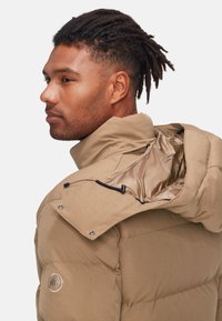 Giacca puffer color khaki con colletto alto, bottoni a pressione e rivestimento interno. Presenta una cerniera sulla spalla e un logo ricamato sul maniche.