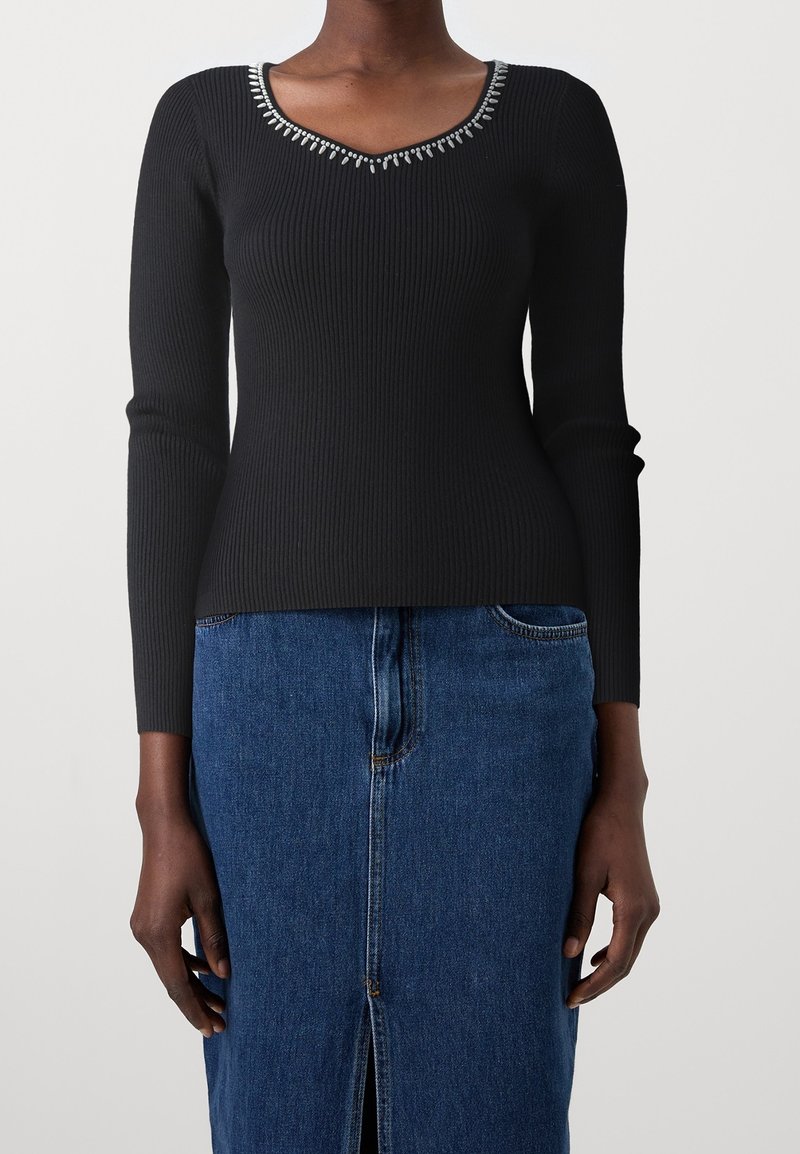 Haut noir à manches longues côtelé avec un col en V orné de détails argentés, porté avec une jupe en denim bleu avec une fente à l'avant.