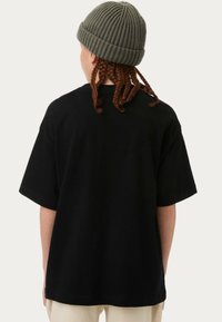 Camiseta negra de gran tamaño con mangas cortas, hecha de algodón. La tela parece suave. La persona lleva un gorro de lana verde y tiene el cabello trenzado.