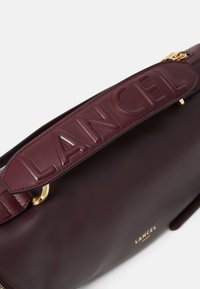 Sac en cuir bordeaux avec une bandoulière texturée estampillée "LANCEL." Quincaillerie dorée et finition lisse, avec fermeture éclair.
