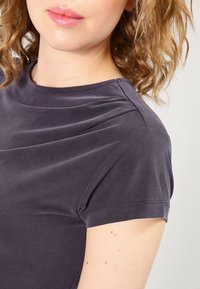 Ciemnoszara bluzka z krótkim rękawem o subtelnej fakturze i podwiniętych rękawach, z okrągłym dekoltem i gładkim wykończeniem z tkaniny.