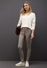 Jeune femme portant un pull blanc, un pantalon ajusté taupe, des sneakers blanches, tenant une veste marron, se tenant devant un fond gris foncé.