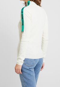Jersey blanco de cuello alto con relieve, que presenta una tira verde con el texto "UNITS OF BENETTON", combinado con jeans azules. Diseño minimalista.
