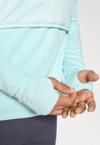 Lyseblå atletisk top med lange ærmer og tommelhuller. Glat stof med en subtil glans, lagdelt design og en kontrasterende pink detalje.