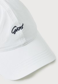 White baseball cap with a curved brim, featuring navy embroidered "Gant" logo. Made from lightweight material with a smooth texture.