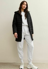 Parka nera con cappuccio, dotata di chiusura con zip e motivo trapuntato verticale, abbinata a pantaloni della tuta grigi chiari e sneakers.