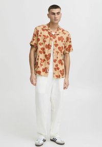 Camisa de manga corta en beige claro con estampado floral rojo, combinada con pantalones de lino blancos y zapatillas grises con acentos negros.