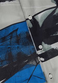 Tkanina koszuli z abstrakcyjnymi wzorami w kolorach czarnym, niebieskim, szarym i białym oraz trzema widocznymi białymi guzikami przyszytymi czarną nicią.