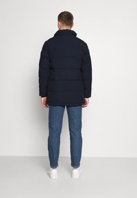 Hombre de pie de espaldas, vistiendo una chaqueta acolchada de color negro marino, jeans azules y zapatillas blancas, frente a un fondo claro y liso.