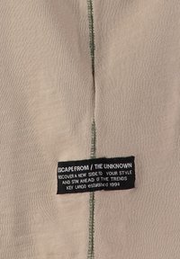 Tessuto beige con cuciture verdi a punto overlock e un'etichetta rettangolare nera con la scritta "Fuggi dall'ignoto, scopri un nuovo lato del tuo stile".