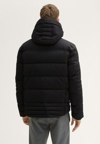 Jachetă puffer neagră cu guler înalt și glugă, având panouri matlasate și o textură netedă, mată. Vedere din spate care arată tivul ajustabil.