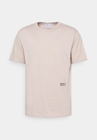 Camiseta de algodón de color beige claro con cuello redondo. Presenta mangas cortas y un pequeño texto de logo negro en la parte inferior izquierda.