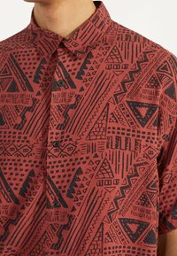 Camisa de manga corta en color óxido con patrones geométricos negros, cuello con botones y un corte relajado. Tejido de algodón texturizado.
