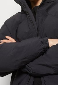 Person klädd i en svart vadderad jacka med dragkedja och elastiska ärmslut, med armarna korsade över bröstet mot en neutral bakgrund.