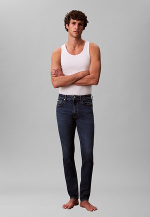 Blugi bleumarin cu croială slim, cusături discrete contrastante, asortați cu un maiou alb. Modelul stă dezbrăcat în picioare, cu brațele încrucișate.