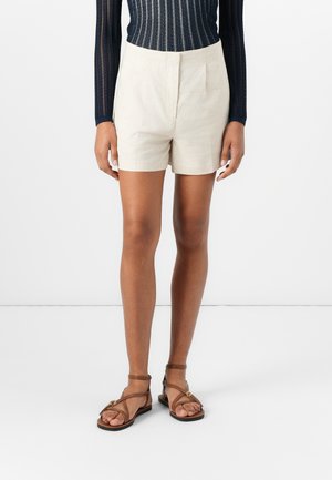 Person iført beige shorts med høj talje, mørkeblå bluse med lange ærmer og brune flade sandaler med remme, stående mod en hvid baggrund.
