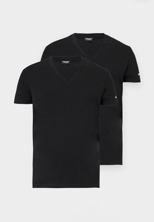 Άνδρας που στέκεται ξυπόλυτος, φοράει μαύρο T-shirt με V-λαιμό και μαύρα στενά σορτς μπροστά σε ένα απλό ανοιχτό γκρι φόντο.