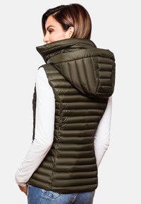 Gilet rembourré vert olive avec un col haut et une capuche, présentant une texture matelassée et une coupe slim, port�é par-dessus une chemise blanche à manches longues.