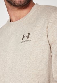 Sudadera beige hecha de tela suave, con cuello redondo y un pequeño logo oscuro en el lado izquierdo del pecho. La textura parece suave y casual.