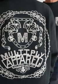 Čierna mikina s bielym vyšívaným dizajnom, dekoratívnymi prvkami, textom "MULTIPLY APPAREL 2025-GER" a stilizovaným písmenom "M."