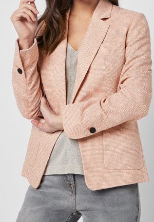 Femme portant un blazer en tweed rose avec des boutons noirs sur un pull en V gris et un jean gris, bras croisés à la taille.