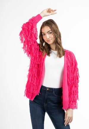 Cardigan fucsia con dettagli in fringes lungo le maniche, abbinato a una maglietta bianca e jeans blu scuro. Il tessuto è testurizzato e leggero.