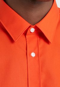 Een persoon met een donkere huid die een feloranje overhemd met kraag draagt, tot aan de bovenkant dichtgeknoopt waarbij de witte knopen zichtbaar zijn.