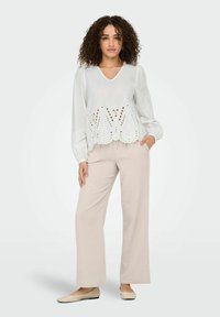Weißes Bluse mit Ausschnitten am Saum, kombiniert mit beigen weit geschnittenen Hosen und hellen Ballerinas. Das Model steht vor einem neutralen Hintergrund.