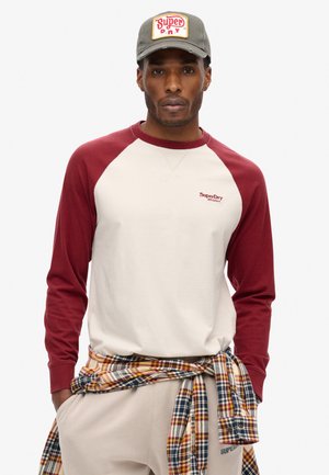 Langsleeve honkbalshirt met een crèmekleurig lichaam en bordeauxrode raglanmouwen. Borduursel op de borst. Gecombineerd met een geruite flanellen shirt om de taille.