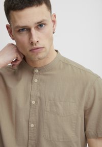 Beige krage skjorte med korte ermer laget av lett stoff, med knapplukking foran og en brystlomme med en subtil tekstur.