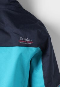 Veste technique bleu marine et turquoise avec une texture de tissu lisse, ornée d'une broderie de logo sur l'épaule et de panneaux de couleur contrastante.