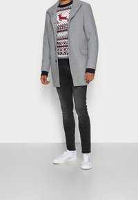 Grå rock i ullblandning med krage, bärs över en mönstrad röd, vit och marinblå tröja, kombinerad med svarta jeans och vita sneakers.