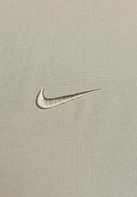 Lichtgrijze stof met een geborduurd gouden Nike-swoosh-logo rechtsonder. De textuur lijkt glad en geweven.
