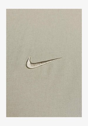 Lys beige stoff har en glatt tekstur med en hevet, brodert Nike swoosh-logo i en litt mørkere beige nyanse.