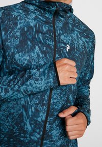 Veste zippée à motif bleu avec capuche ; fabriquée en matériau lisse et extensible ; dotée d'une fermeture éclair noire et d'accents aux poignets.