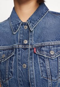 Jeansjacka med klassisk krage, framfickor, silverfärgade knappar och röd etikett, med en texturerad blått tyg och stygn detaljer.