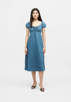 Γυναίκα στέκεται σε λευκό φόντο, φοράει μπλε τζιν midi φόρεμα με φουσκωτά μανίκια και μαύρα πέδιλα με τακούνι.