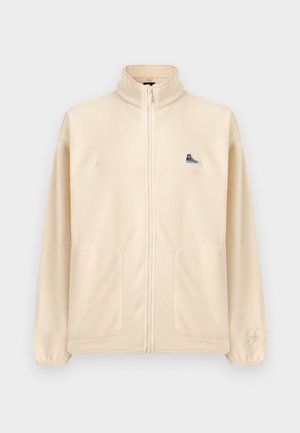 Beige Fleecejacke mit Reißverschluss, hohem Kragen, zwei Fronttaschen, kleinem schwarzen Sneaker-Patch auf der Brust und dezentem Converse-Logo am linken Ärmel.
