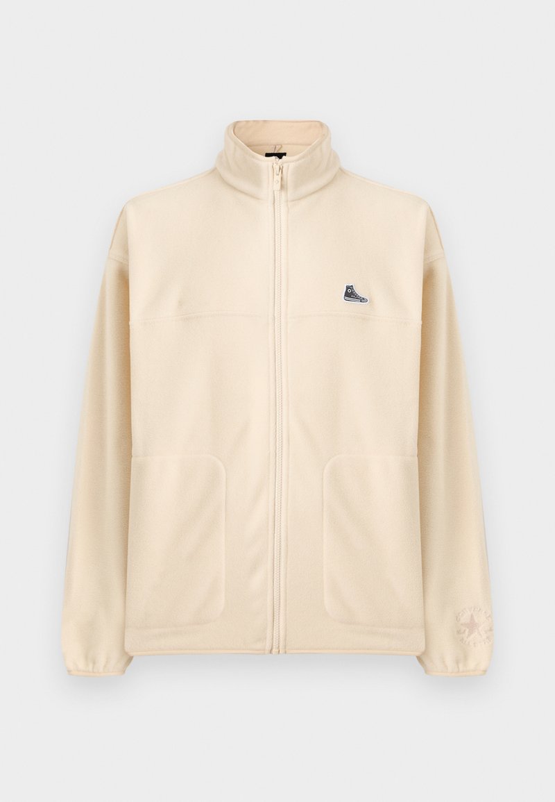 Giacca in pile beige con zip, colletto alto, due tasche frontali, piccola toppa nera a forma di sneaker sul petto e logo sottile Converse sulla manica sinistra.