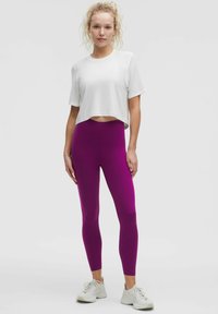 Biela skrátená atletická tričká s textúrovanými rukávmi, v kombinácii s vysokými fialovými legínami. Športová obuv dopĺňa celý outfit.