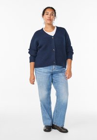 Jūros jėgos mėlynas megztinis su V formos kaklu, keturiais mygtukais ir suktomis rankovėmis, derinamas su šviesiai mėlynomis tiesiomis kelnėmis ir juodais batu.