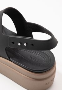 Schwarze Gummisandale mit einer dicken braunen Sohle, ausgestattet mit einem verstellbaren Riemen und silbernen Hardware-Details, strukturiertes Fußbett für besseren Halt.