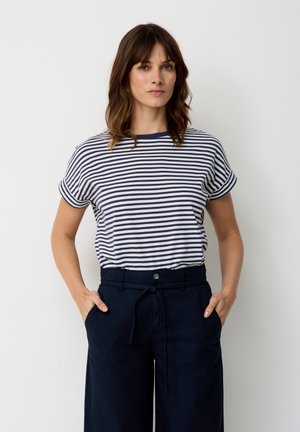 Frau mit schulterlangem braunem Haar, die ein marineblau-weiß gestreiftes kurzärmeliges Shirt und marineblaue Hose mit Kordelzug trägt und mit den Händen in den Taschen steht.